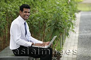 Asia Images Group - Indian man sitting on park bench working on laptop and smiling.