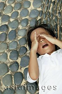 Asia Images Group - Young boy with hands over eyes, lying down on pebbled floor