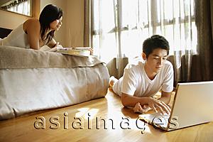 Asia Images Group - Young man using laptop, young woman lying on bed reading magazine