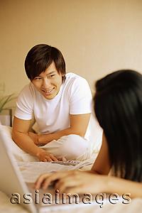 Asia Images Group - Couple in bedroom, woman using laptop and looking at man behind her.