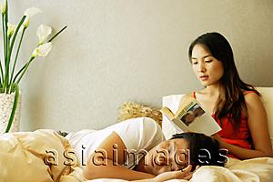 Asia Images Group - Woman on bed, reading a book, man lying on her lap