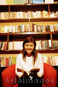 Asia Images Group - Young woman, sitting on chair, reading book, bookshelf behind her
