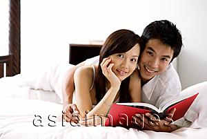 Asia Images Group - Couple lying on bed, holding book, smiling at camera