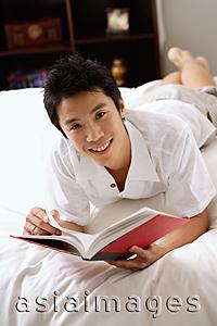 Asia Images Group - Man lying on bed, holding book, smiling at camera