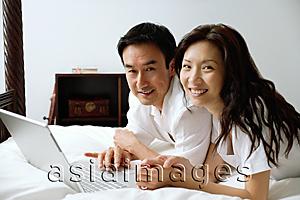 Asia Images Group - Couple lying on bed, using laptop, smiling at camera