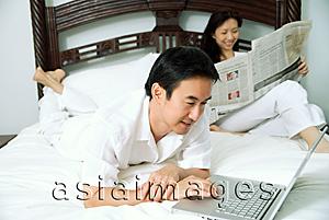 Asia Images Group - Couple in bedroom, man using laptop, woman reading newspaper