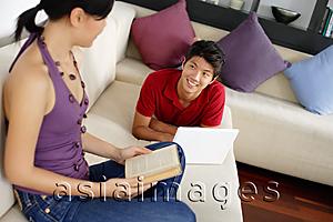 Asia Images Group - Couple in living room, man lying on sofa with laptop, woman holding book