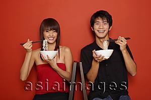 Asia Images Group - Couple sitting against red wall, holding bowl of noodles, smiling at camera