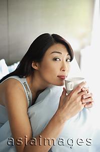 Asia Images Group - Woman sitting on bed, holding cup to lips, looking at camera