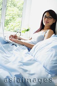 Asia Images Group - Woman sitting up on bed with laptop, looking at camera