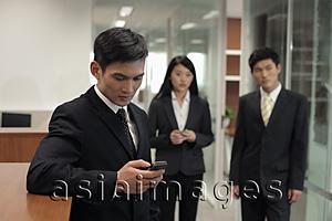 Asia Images Group - Three businesspeople standing in office, one texting on a phone