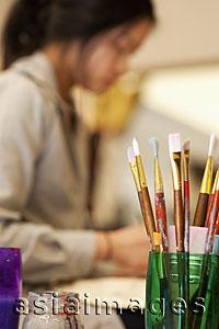 Asia Images Group - Paint brushes in the foreground with young woman painting in the background