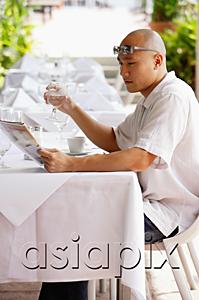 AsiaPix - Man sitting at table, reading newspaper, holding glass of water