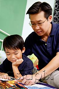 AsiaPix - Boy lying on bed, reading a book, father next to him pointing at book