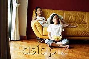 AsiaPix - Two girls sitting in living room, one holding remote control, the other holding bowl of popcorn