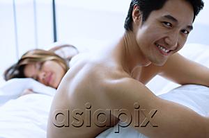 AsiaPix - Man sitting on bed, smiling at camera, woman asleep next to him