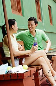 AsiaPix - Couple sitting on bench on tennis court, smiling at each other, woman with legs crossed