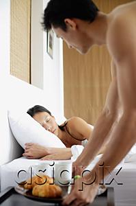 AsiaPix - Woman sleeping in bed, man setting down breakfast tray next to her on bedside table