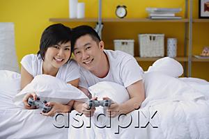 AsiaPix - Couple lying on bed, holding video game controls, smiling at camera
