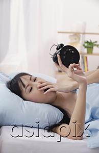 AsiaPix - Young woman lying on bed, looking at alarm clock, yawning