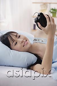 AsiaPix - Young woman lying on bed, looking at alarm clock, smiling