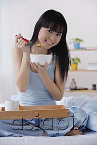 AsiaPix - Young woman sitting on bed, having breakfast, smiling at camera