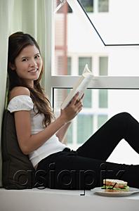 AsiaPix - Woman sitting on bay window holding a book, smiling at camera