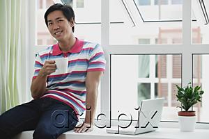 AsiaPix - Man sitting on bay window, holding a cup, laptop open next to him