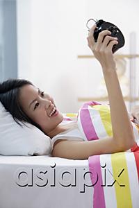 AsiaPix - Young woman lying on bed, looking at alarm clock, smiling