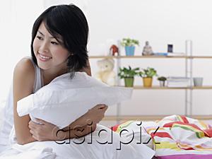 AsiaPix - Young woman sitting on bed, hugging pillow, looking away