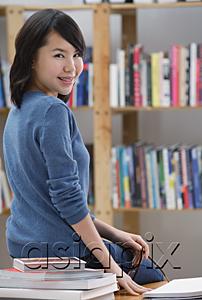 AsiaPix - Young woman in library, sitting on table, looking over shoulder at camera