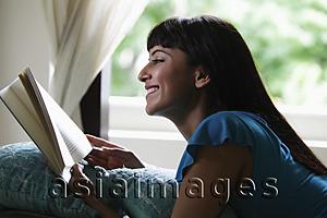 Asia Images Group - Young woman laying on stomach reading a book and smiling