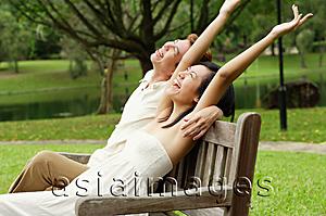 Asia Images Group - Couple sitting on park bench, looking up, woman with arms outstretched
